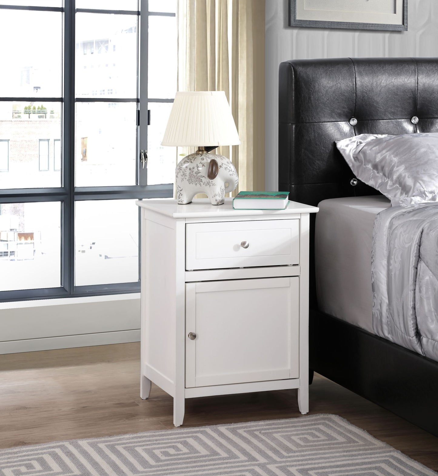 The Izzy 1 Drawer/1 Door Nightstand sits beside a black upholstered bed, topped with a white lamp, a decorative elephant, and a book. Large windows and beige curtains fill the modern bedroom with light.