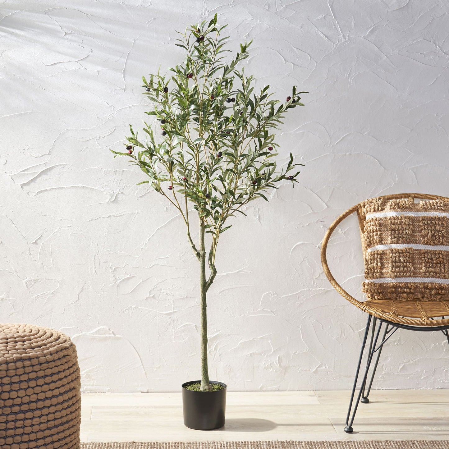 An Artificial Olive Tree with green leaves and dark olives stands on a light floor beside a round woven ottoman and wicker chair, set against a textured white wall.