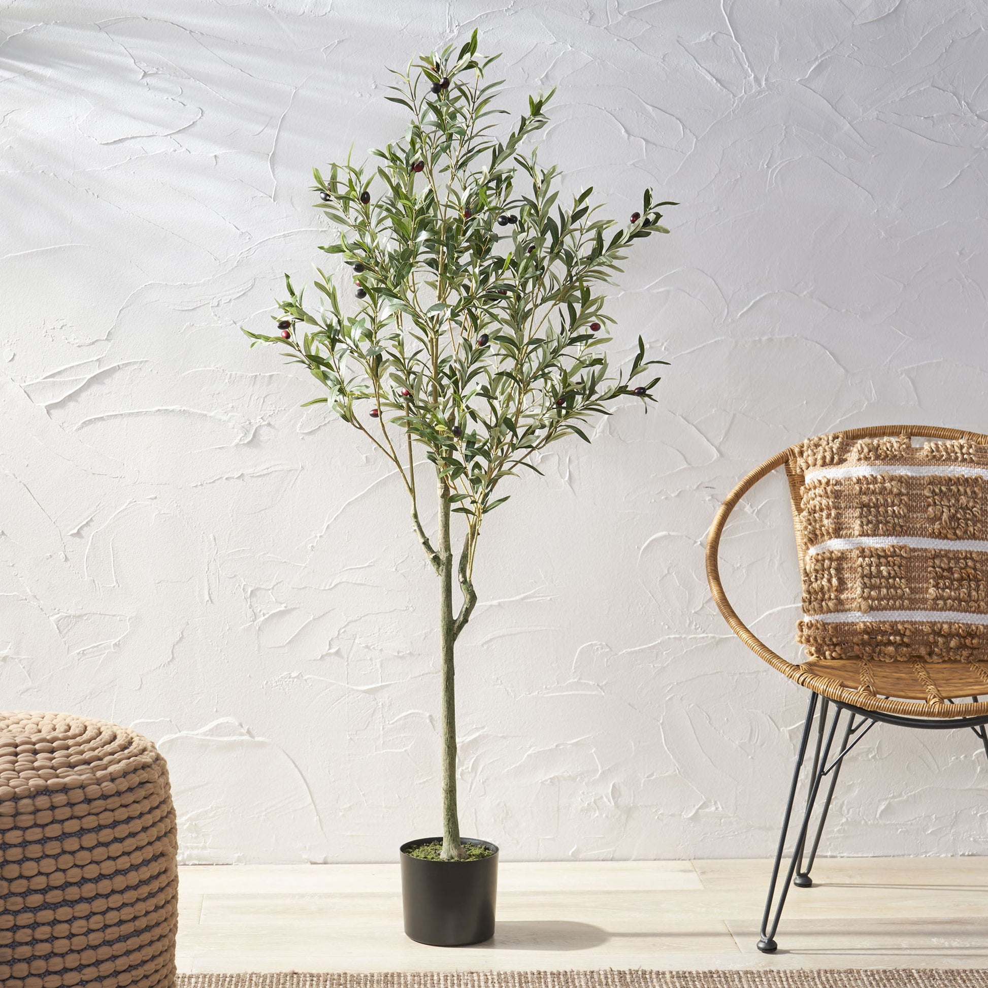 An Artificial Olive Tree with green leaves and dark olives stands on a light floor beside a round woven ottoman and wicker chair, set against a textured white wall.
