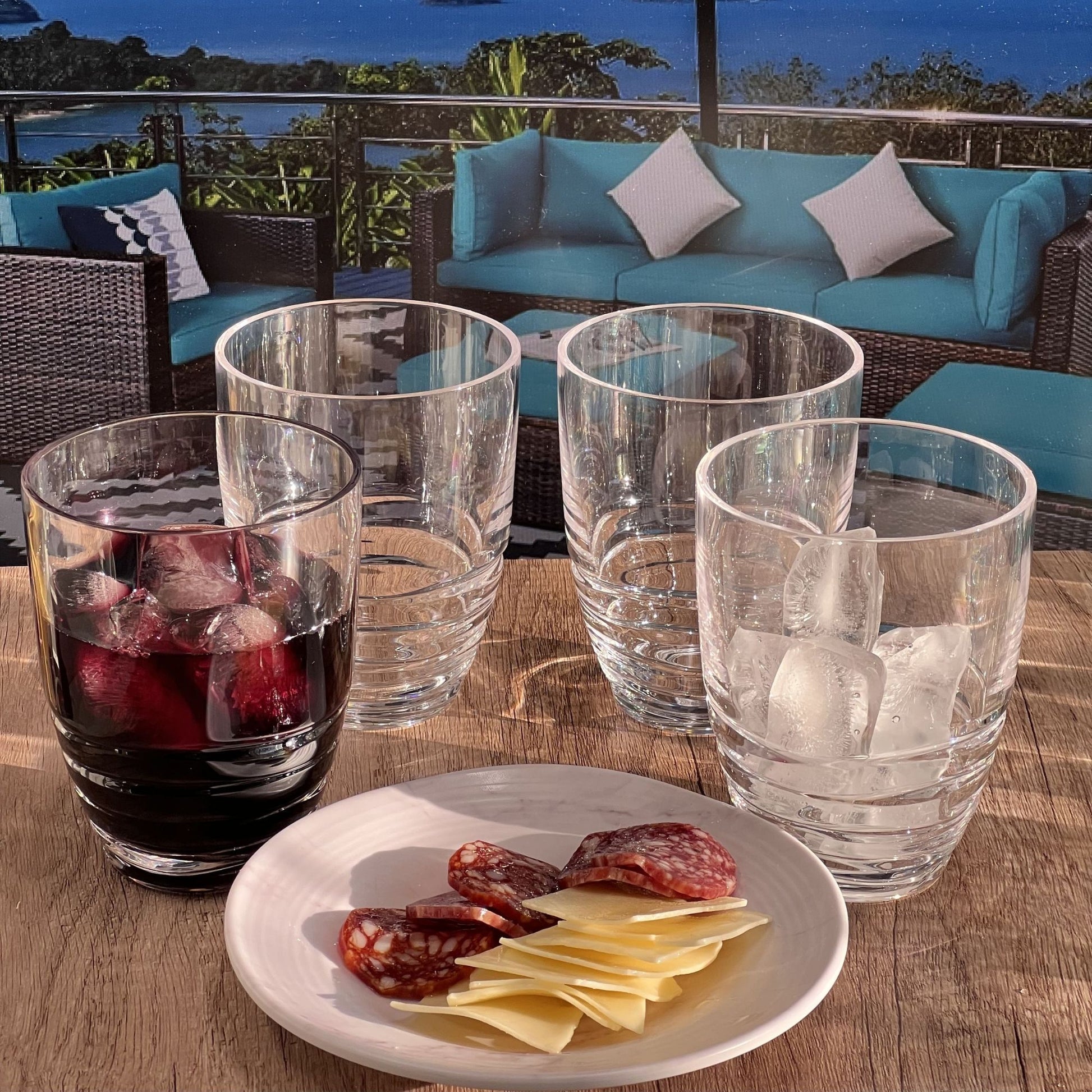 A set of Leading Ware Swirl Acrylic Drinking Glasses (Set of 4)—two empty, one with a red drink and ice, one with just ice—sits on a table alongside sliced cheese and salami, with an outdoor patio set and scenic view in the background.