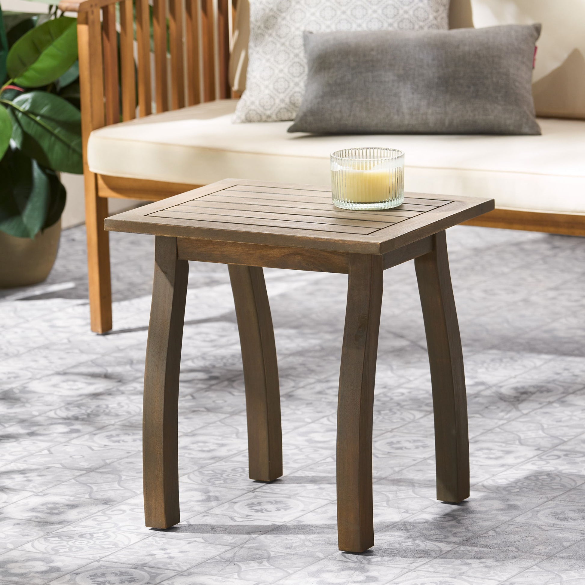The Kinesis Outdoor Side Table with a slatted top and wood frame holds a glass candle and sits on a tile floor beside a cushioned outdoor bench and potted plant.