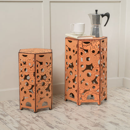 The Selva - Accent Table Set, featuring two decorative orange nested tables with intricate cut-out designs, is placed on a light wood floor near a white paneled wall. The taller table displays a coffee maker, mug, and book.