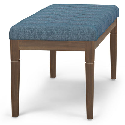 The Waverly Upholstered Tufted Ottoman Bench features a cushioned, textured blue seat with tufted detailing and four straight wooden legs. Displayed against a plain white background.