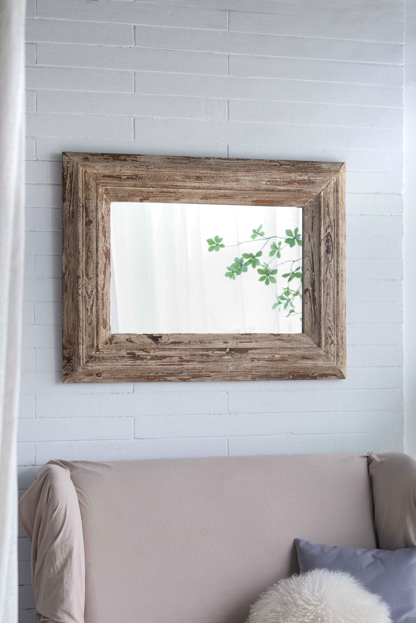 A Rectangle Wall Accent Mirror With Distressed Wood Frame - Brown hangs on a white brick wall above a beige sofa with a gray pillow and fluffy white cushion, reflecting green leaves and soft natural light.