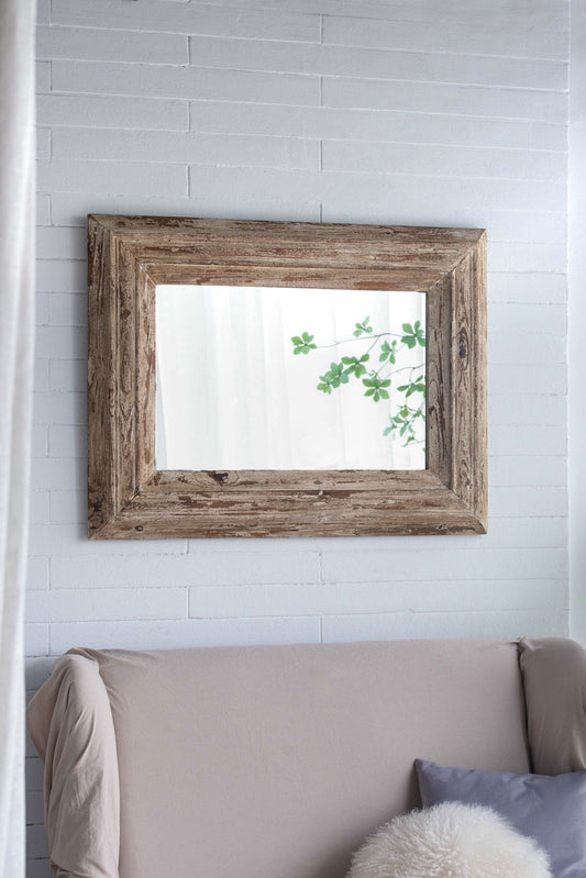 A Rectangle Wall Accent Mirror With Distressed Wood Frame - Brown hangs on a white brick wall above a beige sofa with a gray pillow and fluffy white cushion, reflecting green leaves and soft natural light.