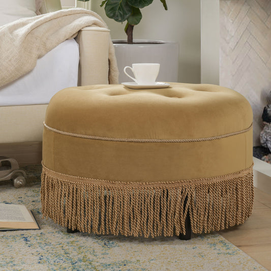 The Yolanda - Luxurious Upholstery Round Upholstered Accent Ottoman in gold velvet with fringe stands on a rug near a beige sofa, topped with a white teacup and saucer, while an open book and potted plant add cozy touches nearby.