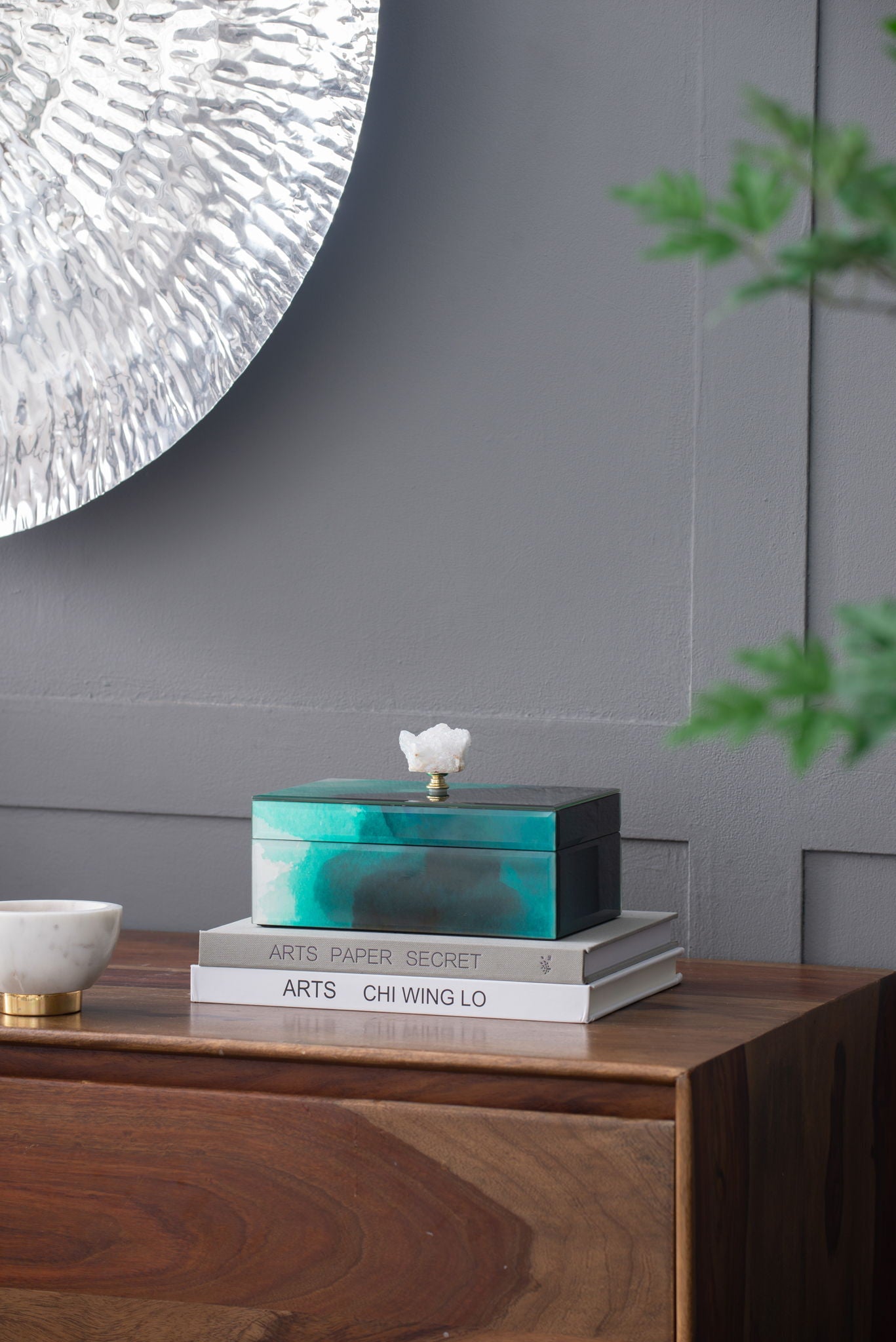 The Bethany - Aqua Jewelry Box is stacked on art books atop a wooden side table, with a marble bowl nearby and silver wall decor above. A green plant peeks in from the right.