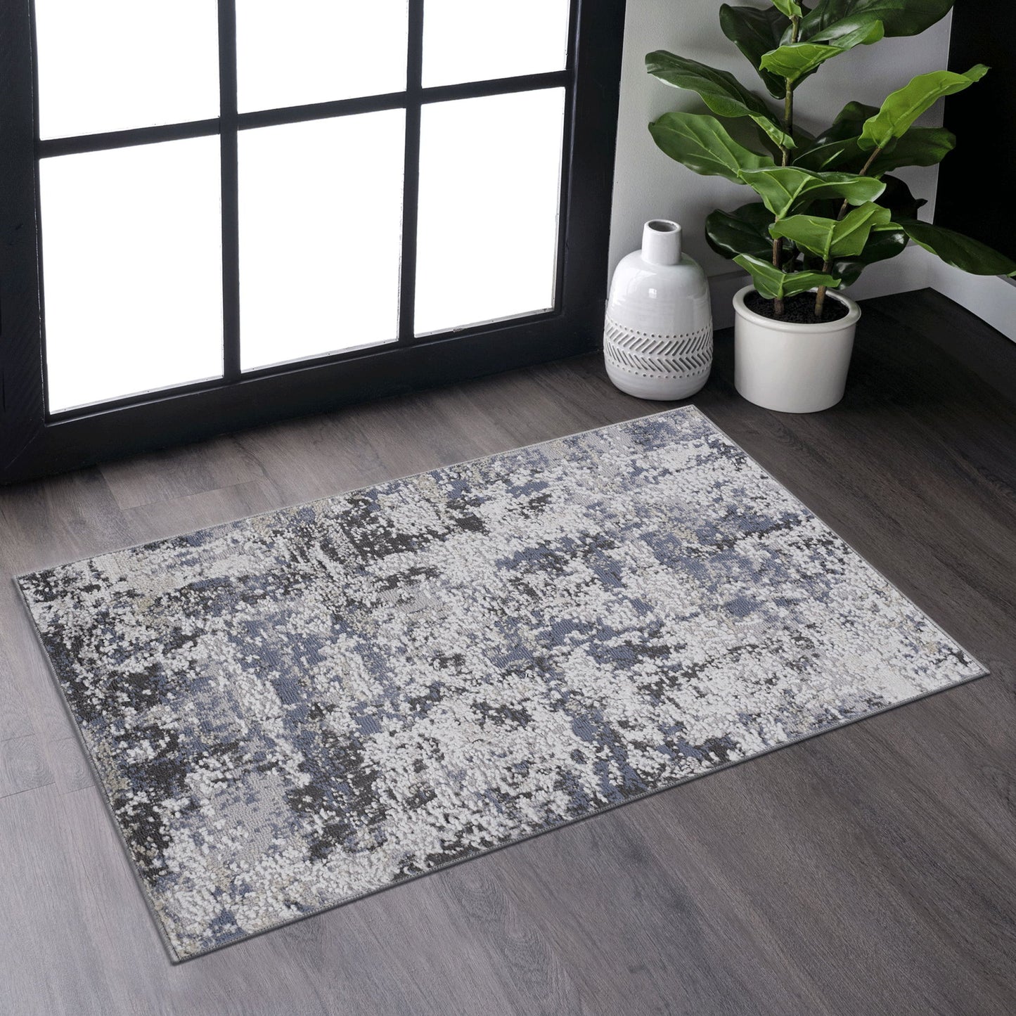 The Payas - Abstract Area Rug, featuring modern blue, gray, and white patterns, rests on a dark wood floor by a large window. Nearby are a potted plant and a white decorative vase.