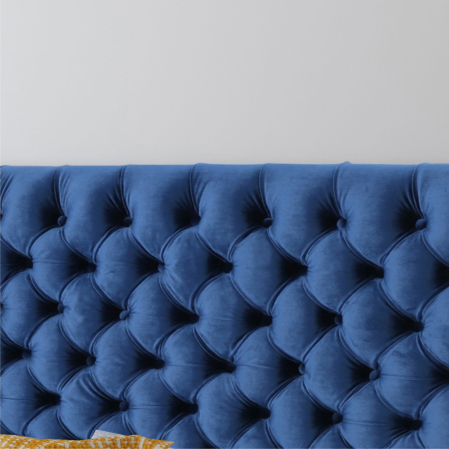 Close-up of the Annecy - Contemporary Upholstered Headboard in blue velvet, tufted design, set against a plain white wall with part of a yellow patterned pillow visible in the bottom left corner.