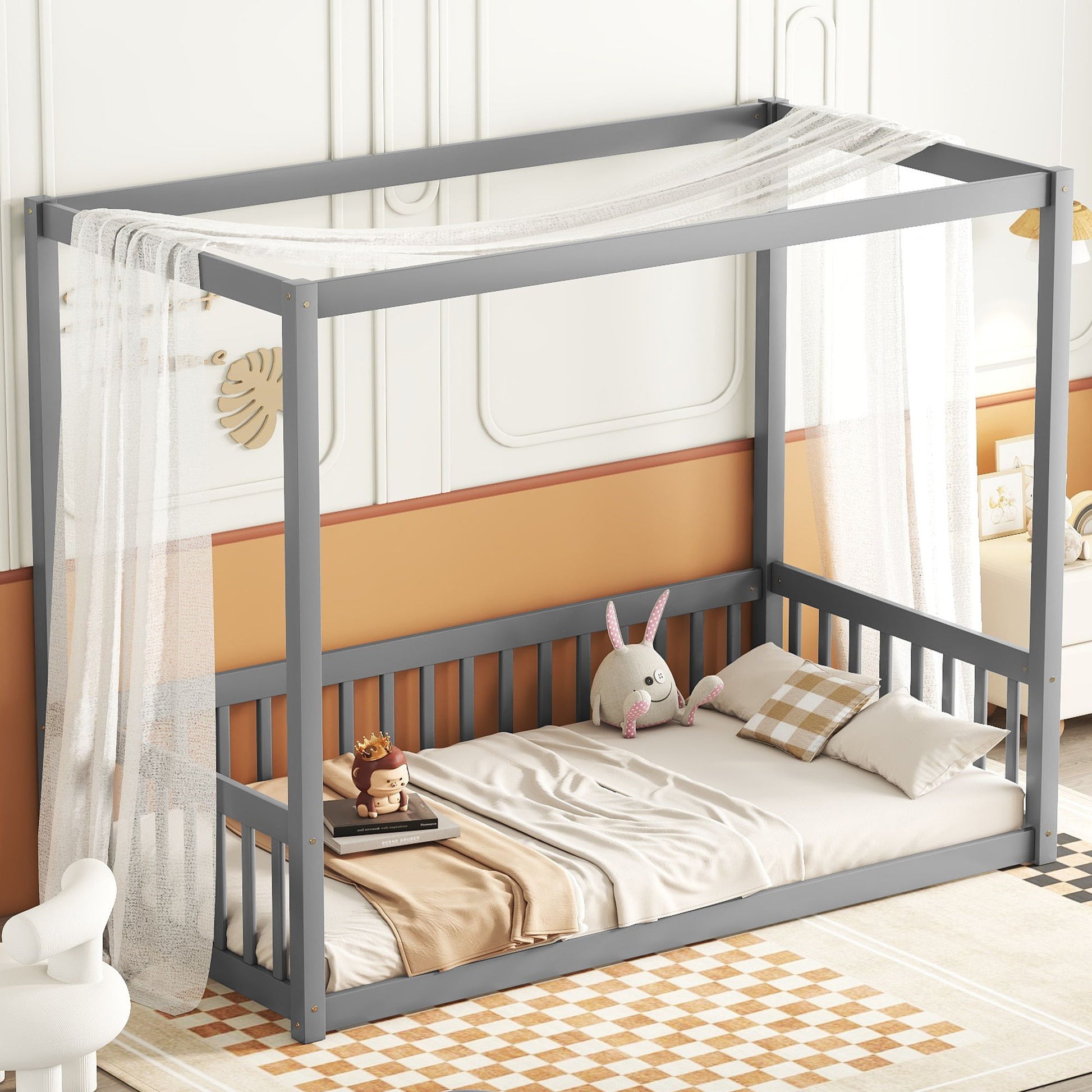 The Canopy Frame Floor Bed With Fence, Guardrails features a gray canopy draped with sheer white fabric, beige bedding, pillows, and stuffed toys. It sits in a warmly decorated room with orange accents and a checkered rug.