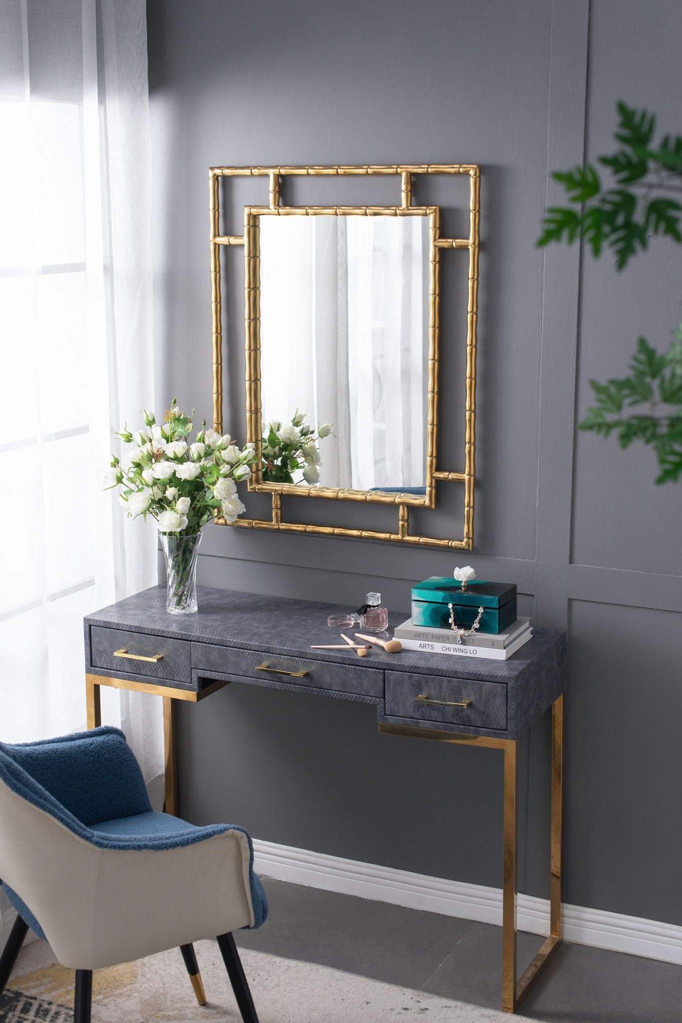 A modern vanity table with gold legs sits against a gray wall. On top are white flowers in a vase, stacked books, and the Bethany - Aqua Jewelry Box. A gold-framed mirror hangs above and a cushioned chair is nearby.