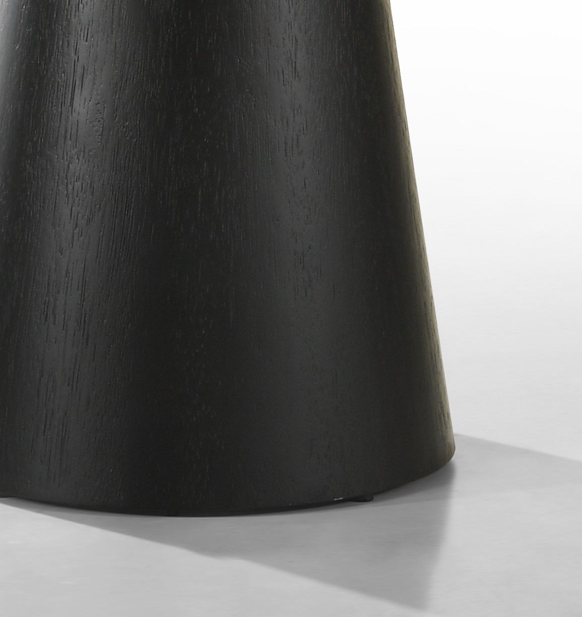 Close-up of the lower portion of the black, cylindrical wooden base of the Jasper - Round Table on a light floor, casting a visible shadow.