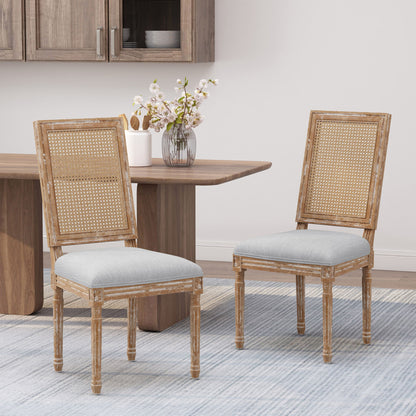 Two French Country Dining Chairs with rectangular cane backrests and smooth upholstered seats are set on a light blue rug in the dining area, near a wooden table with a vase of flowers and a cabinet in the background.