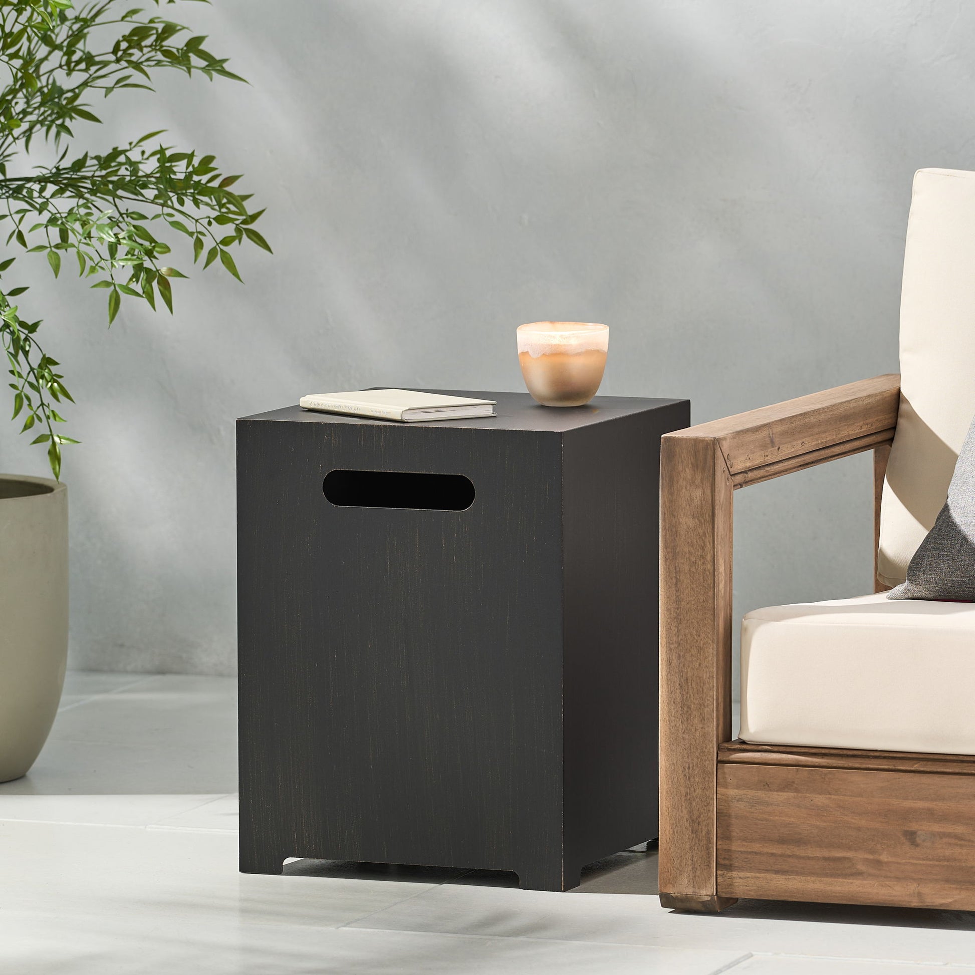 The Modern Tank Holder Side Table With Powder Coated Frame in black holds a closed book and latte beside a wooden armchair with white cushions, near a leafy plant against a light gray wall.