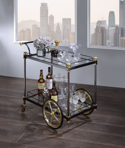 The Cyrus Clear Glass Serving Cart in Black/Gold displays assorted liquor bottles, cocktail glasses, a crystal decanter, and a gray floral arrangement by large windows with city skyline views.