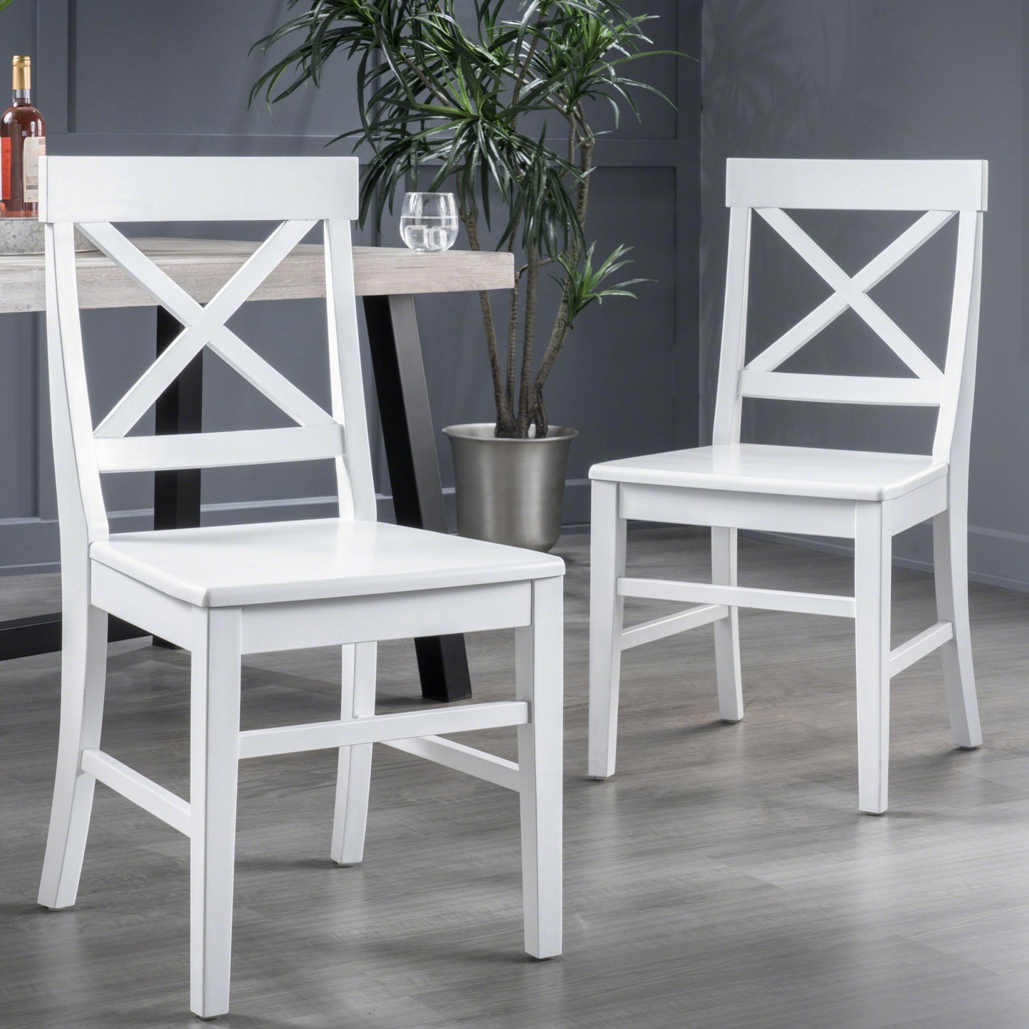 Two Plethora Dining Chairs with X-shaped backs (set of 2) in white sit on a gray floor near a table holding a glass of water and wine bottle, with a potted plant against the gray wall in the background.