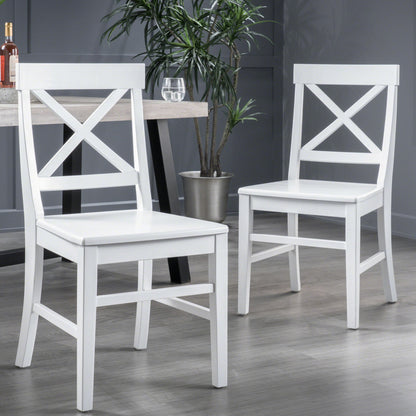 Two Plethora Dining Chairs with X-shaped backs (set of 2) in white sit on a gray floor near a table holding a glass of water and wine bottle, with a potted plant against the gray wall in the background.