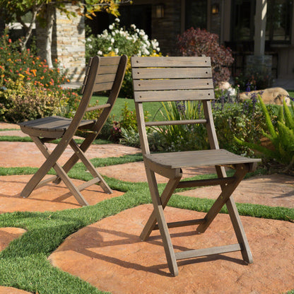 Two Outdoor Foldable Wood Dining Chairs With Slat Design (Set of 2) are set on a stone patio surrounded by green grass and lush garden plants, with a house and blooming flowers in the background.