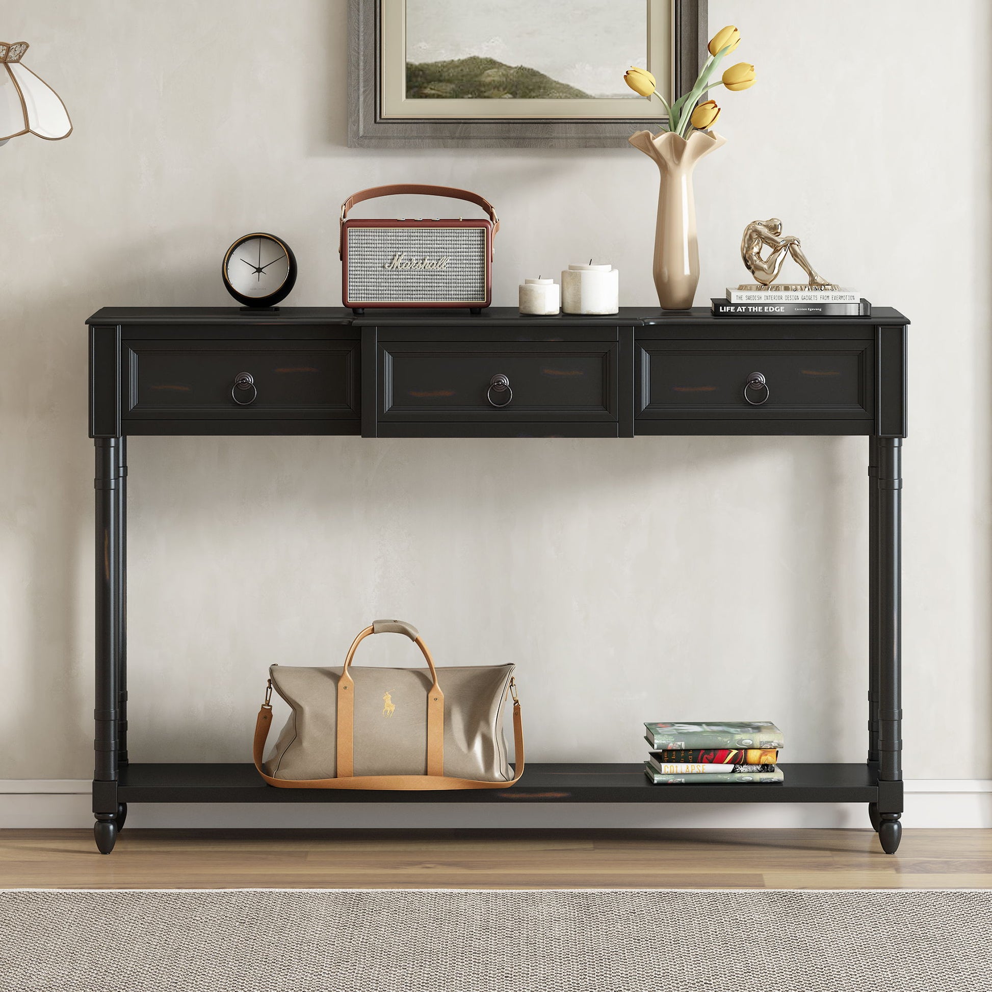 The Console Table Sofa Table With Drawers For Entryway features three drawers holding a vintage radio, a vase with yellow tulips, and books. On the long lower shelf are a beige duffel bag and stacked magazines, with framed artwork displayed above.
