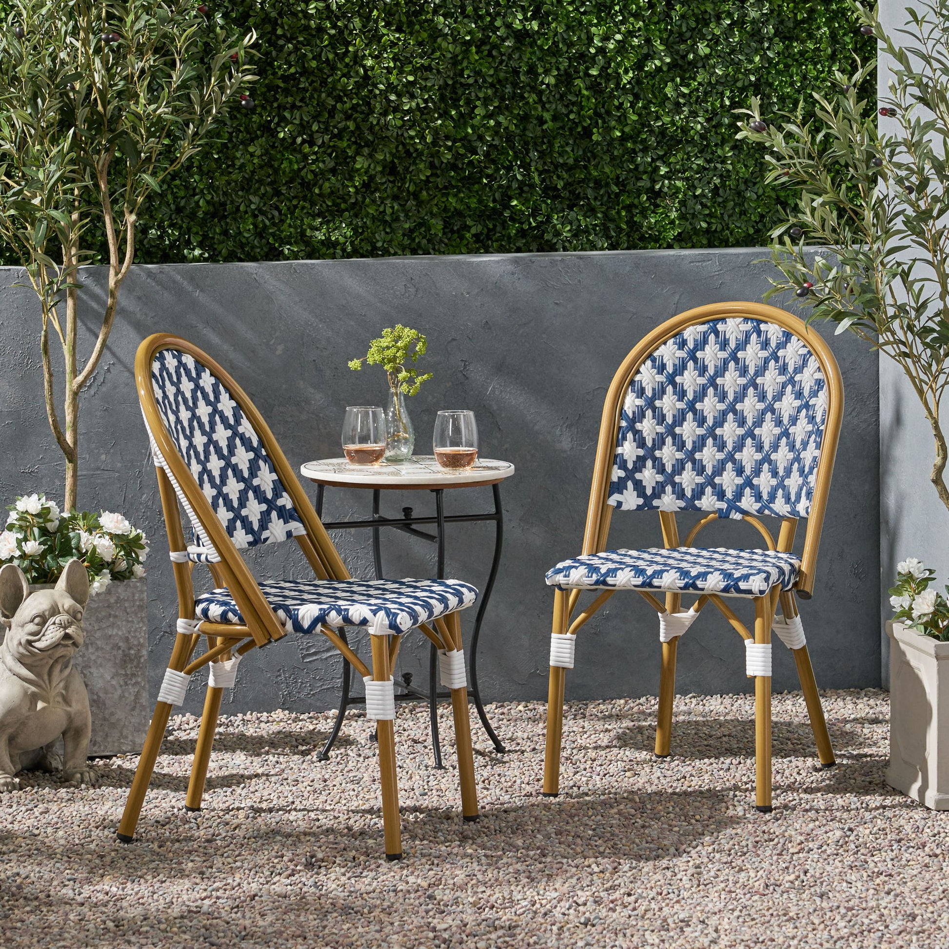 Two Chic French Bistro Chairs with durable rattan and aluminum frames are paired with a small round table holding two glasses on a gravel patio, surrounded by greenery, potted plants, and a decorative dog statue.