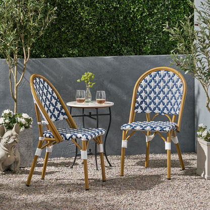 Two Chic French Bistro Chairs with durable rattan and aluminum frames are paired with a small round table holding two glasses on a gravel patio, surrounded by greenery, potted plants, and a decorative dog statue.