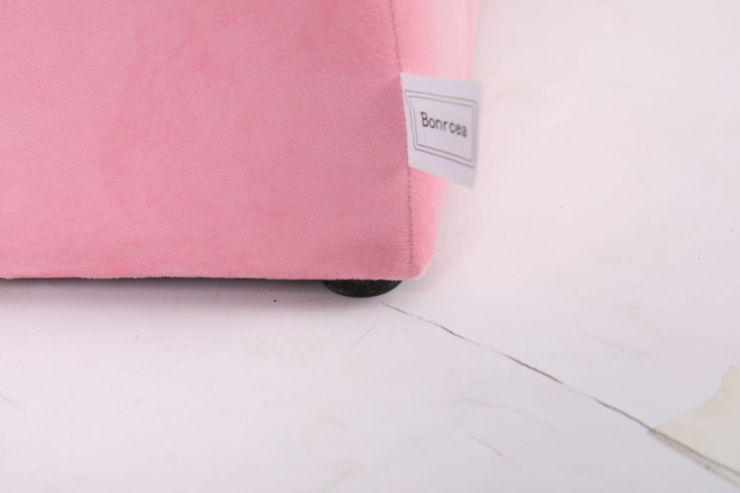 Close-up of the bottom corner of a pink Durable Design Kids Club Sofa, featuring a white rectangular "Bonro" tag, resting on a light, slightly cracked floor.