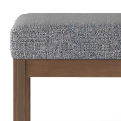 Close-up of the Milltown Upholstered Ottoman Bench, featuring a textured gray cushion and wooden frame, highlighting the fabric and upper part of the legs.
