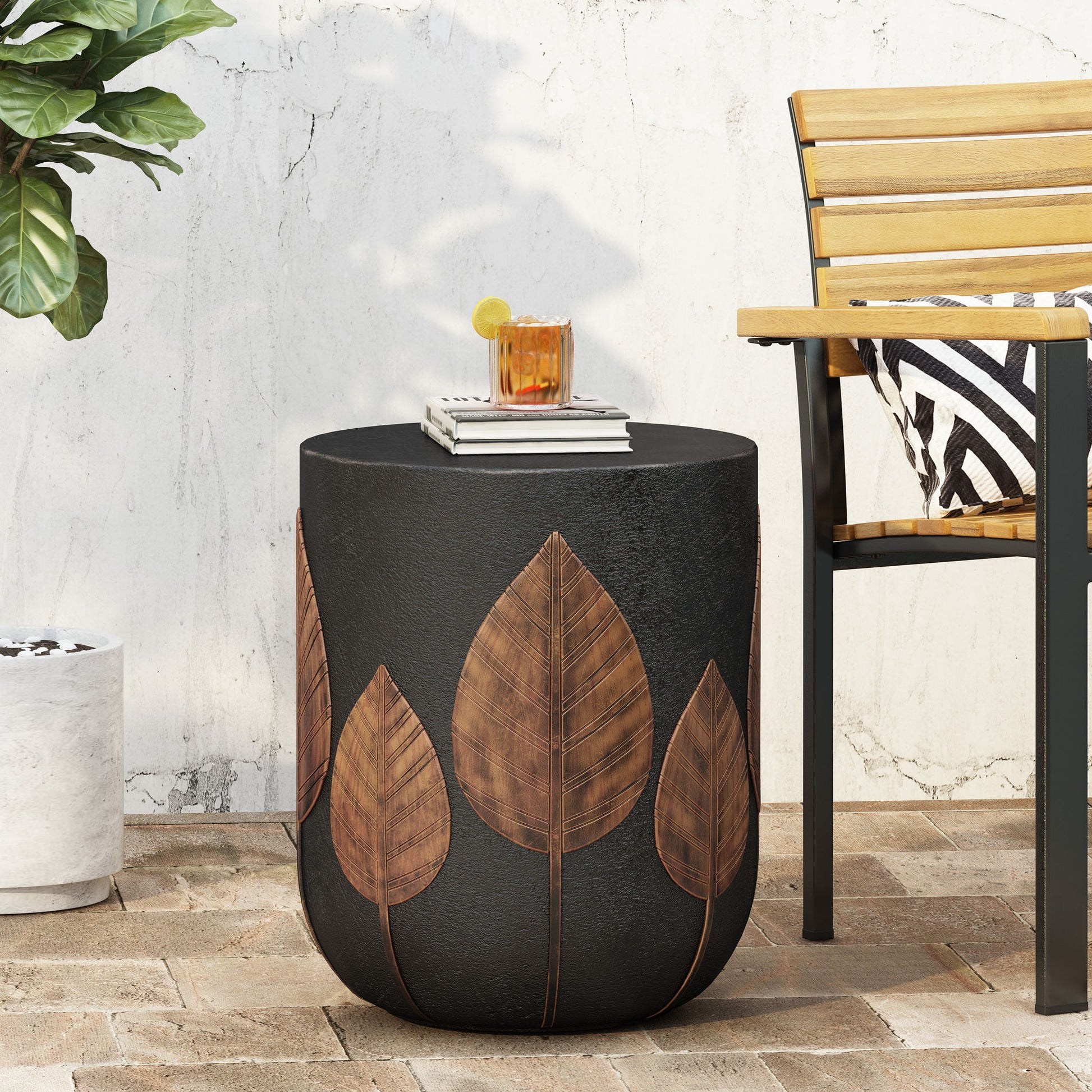 The Outdoor Lightweight Concrete Side Table - Copper, a black cylindrical table with brown leaf designs, sits on the patio next to a wooden chair with a striped cushion, holding a potted plant and a glass of orange drink.