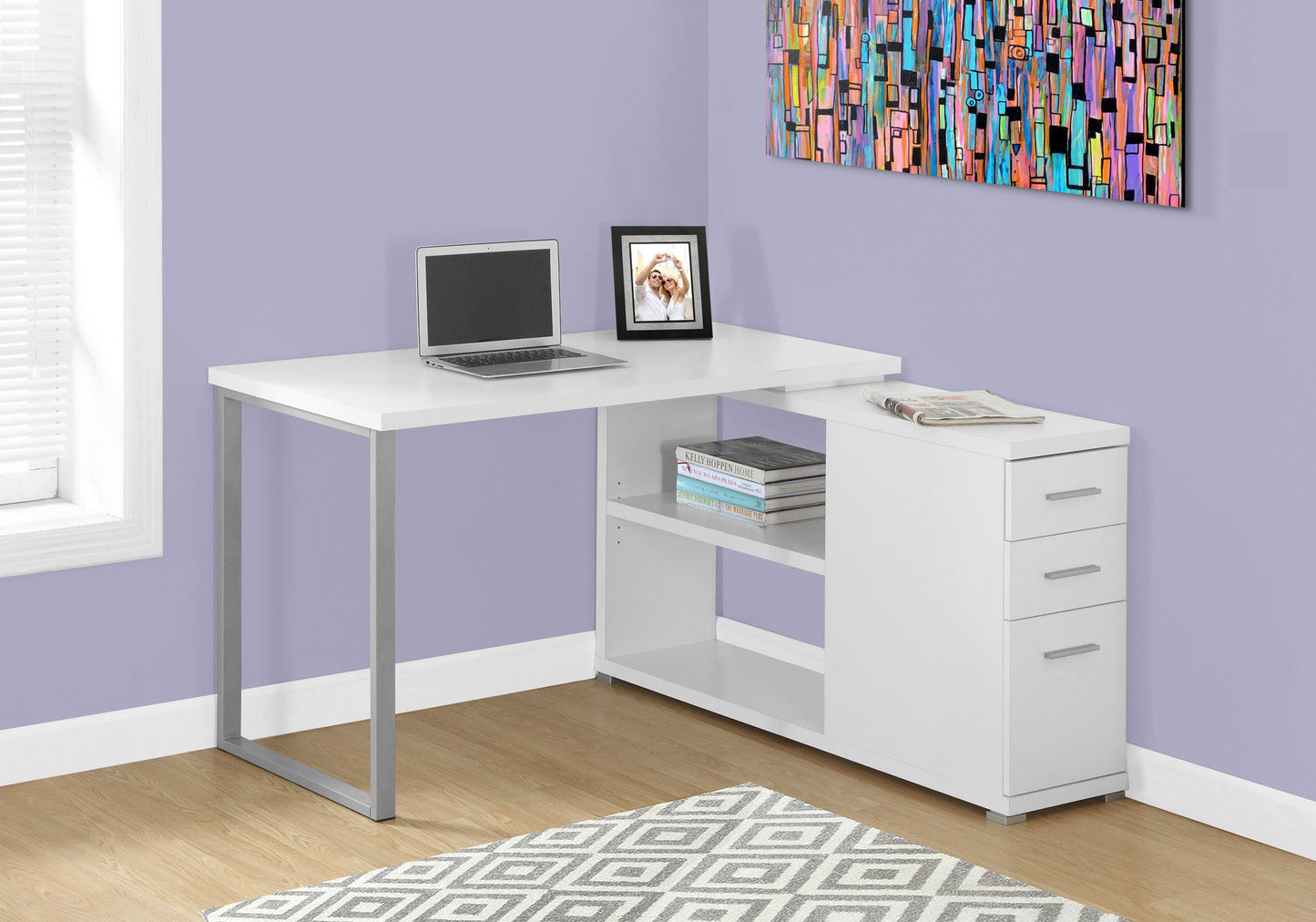 A Computer Desk, Home Office, L Shape with storage drawers and a modern design holds a laptop, books, and décor in a room with purple walls, wood floors, a geometric rug, and colorful wall art.