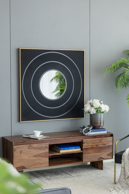 A modern wooden console table with books, a white teacup, and a vase of white flowers sits against a gray wall with the Square Carved Mirror With Pleated Design With Iron Frame in a neutral colorway. Green plants decorate the space.
