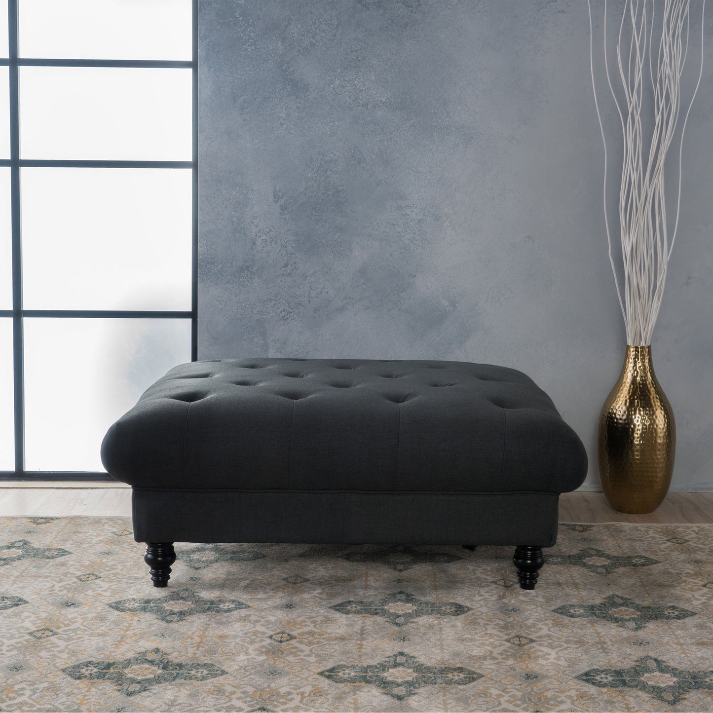 The Madrid - Tufted Ottoman Footrest in dark gray with wooden legs sits on a patterned rug by a textured gray wall, next to a large gold vase with tall white decorative branches. Natural light streams in from the left window.