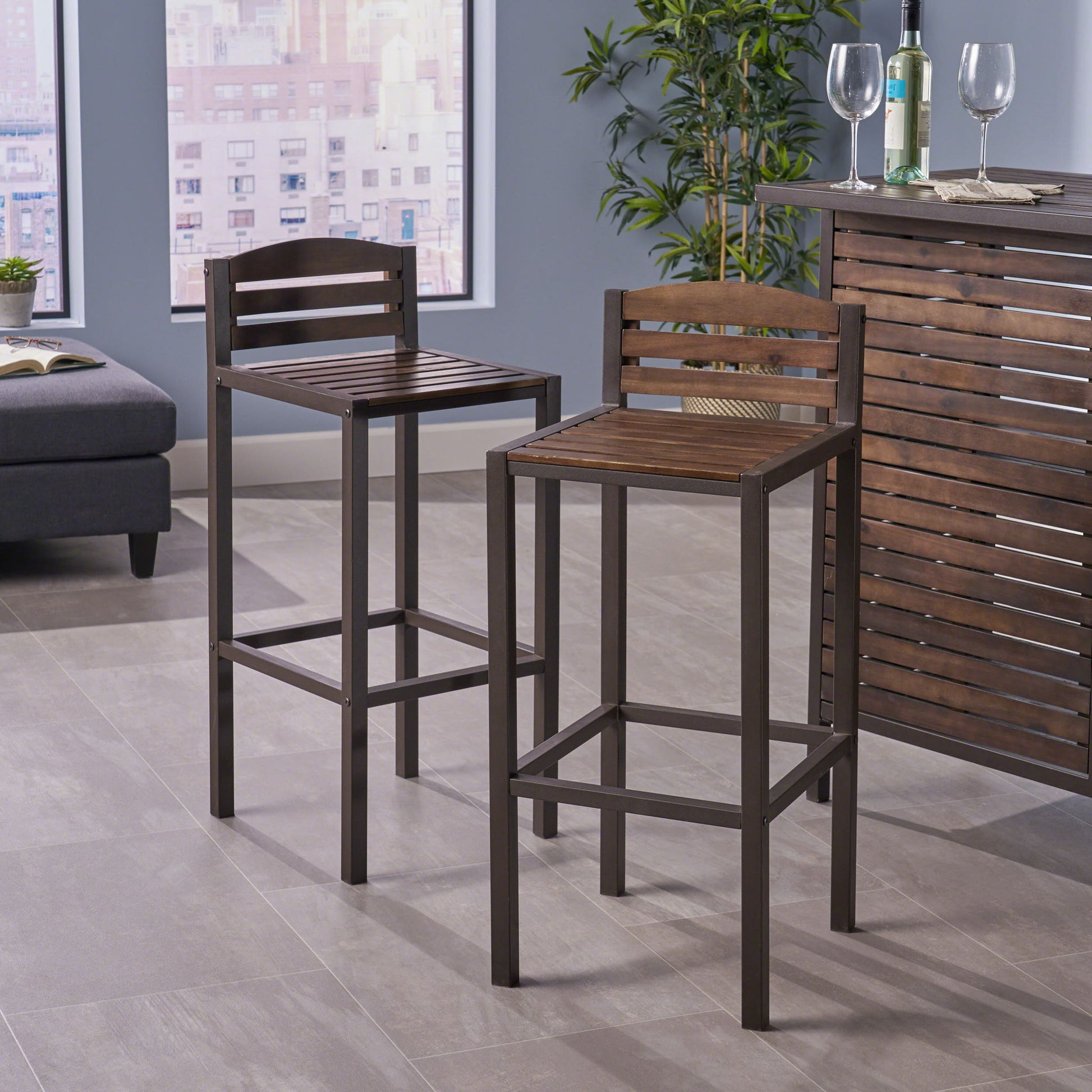Two Outdoor Barstools With Slat Panel Design (Set of 2) in dark brown are placed by a matching wooden bar in a modern room with large windows, a potted plant, wine glasses, and views of city buildings outside.