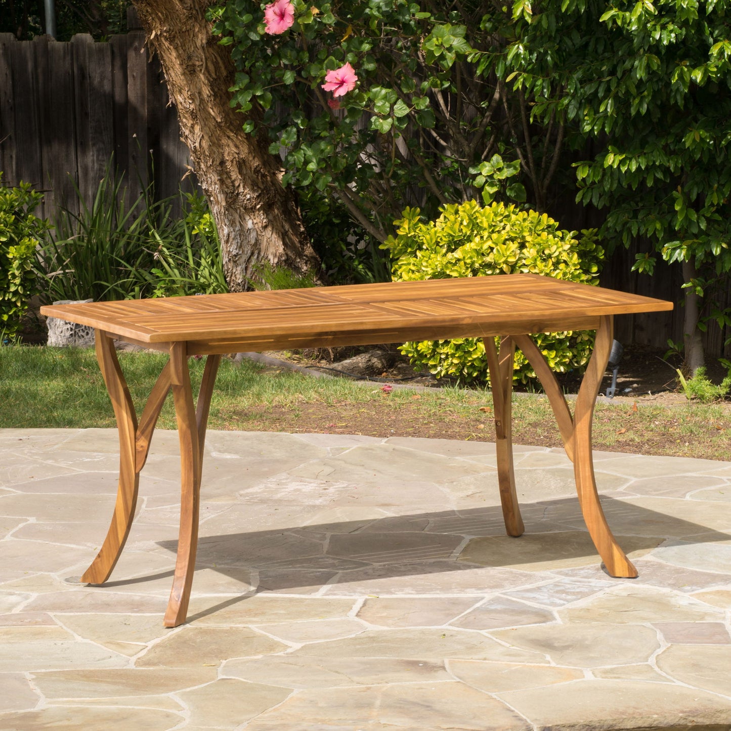 The Outdoor Rectangular Table With Curved Legs And Perpendicular Wooden Slats Table Top stands on a stone patio in the garden, surrounded by green bushes, trees, and blooming pink flowers.