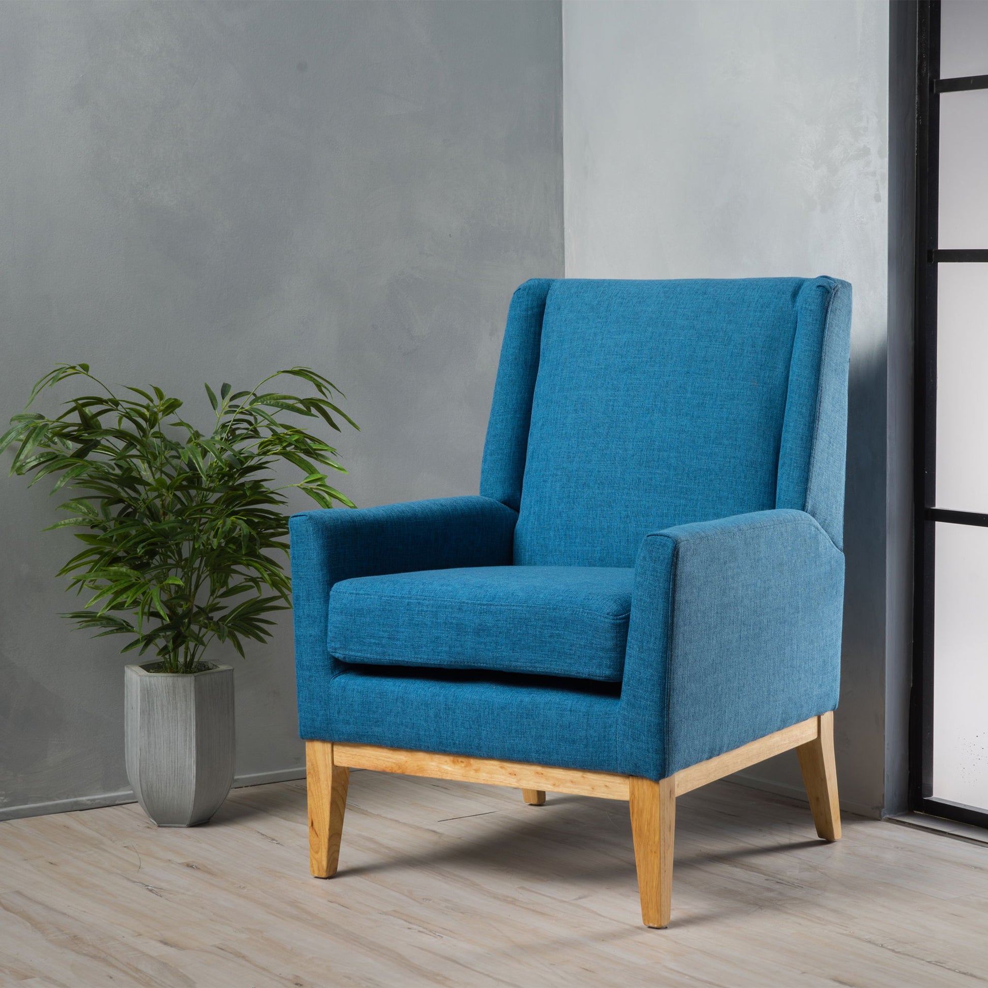 The Modern Accent Chair With Padded Cushion Seat, in blue with wooden legs, is placed on a light wooden floor beside a green potted plant, set against a gray wall near a window with black frame.