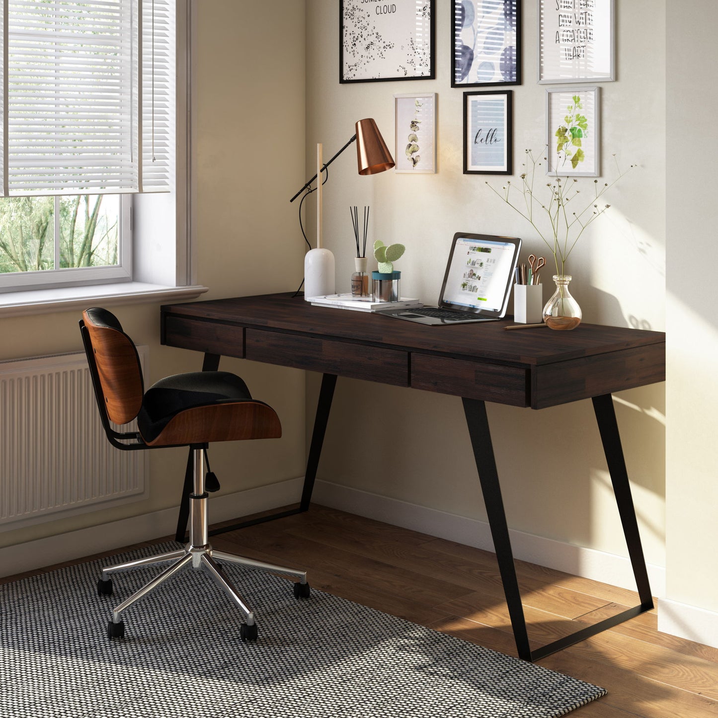 A modern home office showcases the Lowry - Handcrafted Desk with black legs, paired with a swivel chair, laptop, desk lamp, vase with branches, framed wall art, and sunlight shining onto a patterned rug.