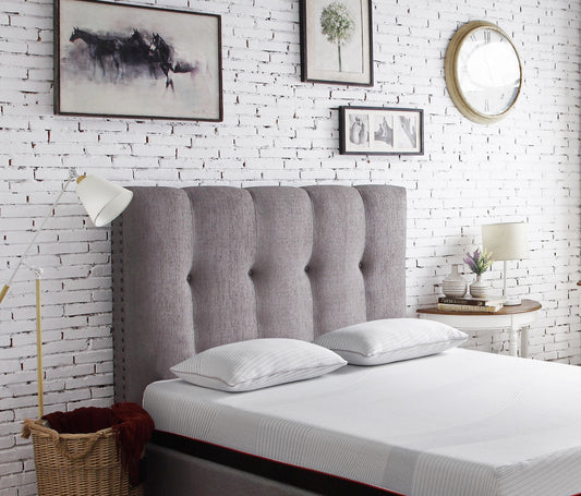A modern bedroom features the Couch.com Queen Size Tufted Upholstered Headboard with nailhead trim in dark gray fabric, two white pillows, a wicker basket, floor lamp, and side table against a white brick wall adorned with art and a round clock.