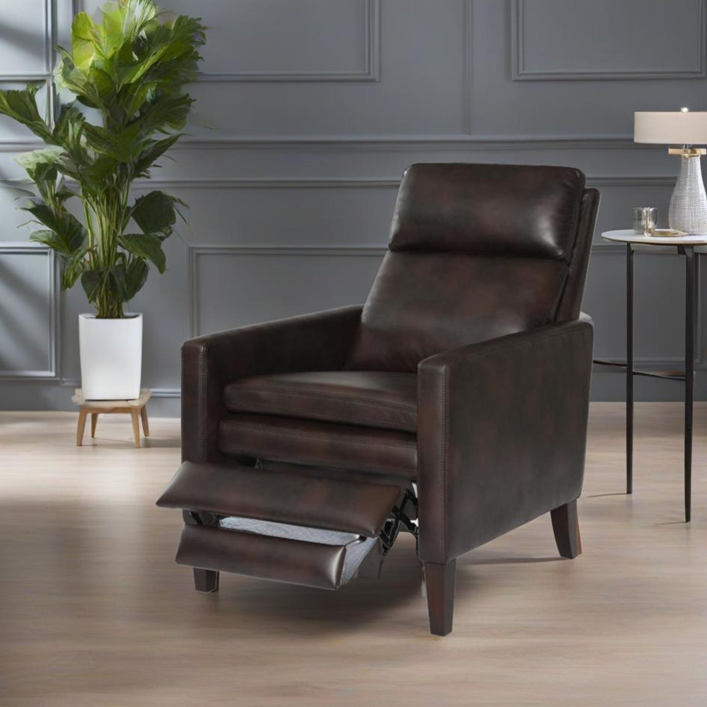 The Vaughn - Push Back Recliner With Solid Hardwood Legs in dark brown leather, featuring an extended footrest, is placed on a light wood floor in a modern room beside a potted plant and a side table with a lamp.