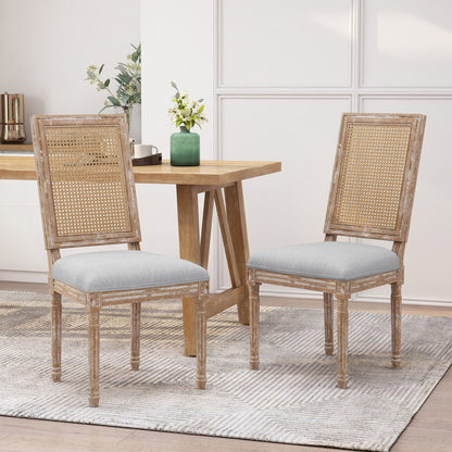 Two French Country Dining Chairs with rectangular cane backrests and smooth upholstered seats sit on a light patterned rug by a wooden dining table. A green vase with flowers is on the table, set against white paneled walls.