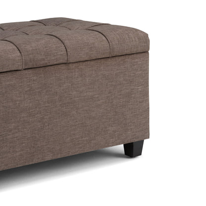 A close-up of the Sienna Upholstered Storage Ottoman Bench featuring brown fabric, a tufted cushioned lid, and short black legs, shown against a white background.