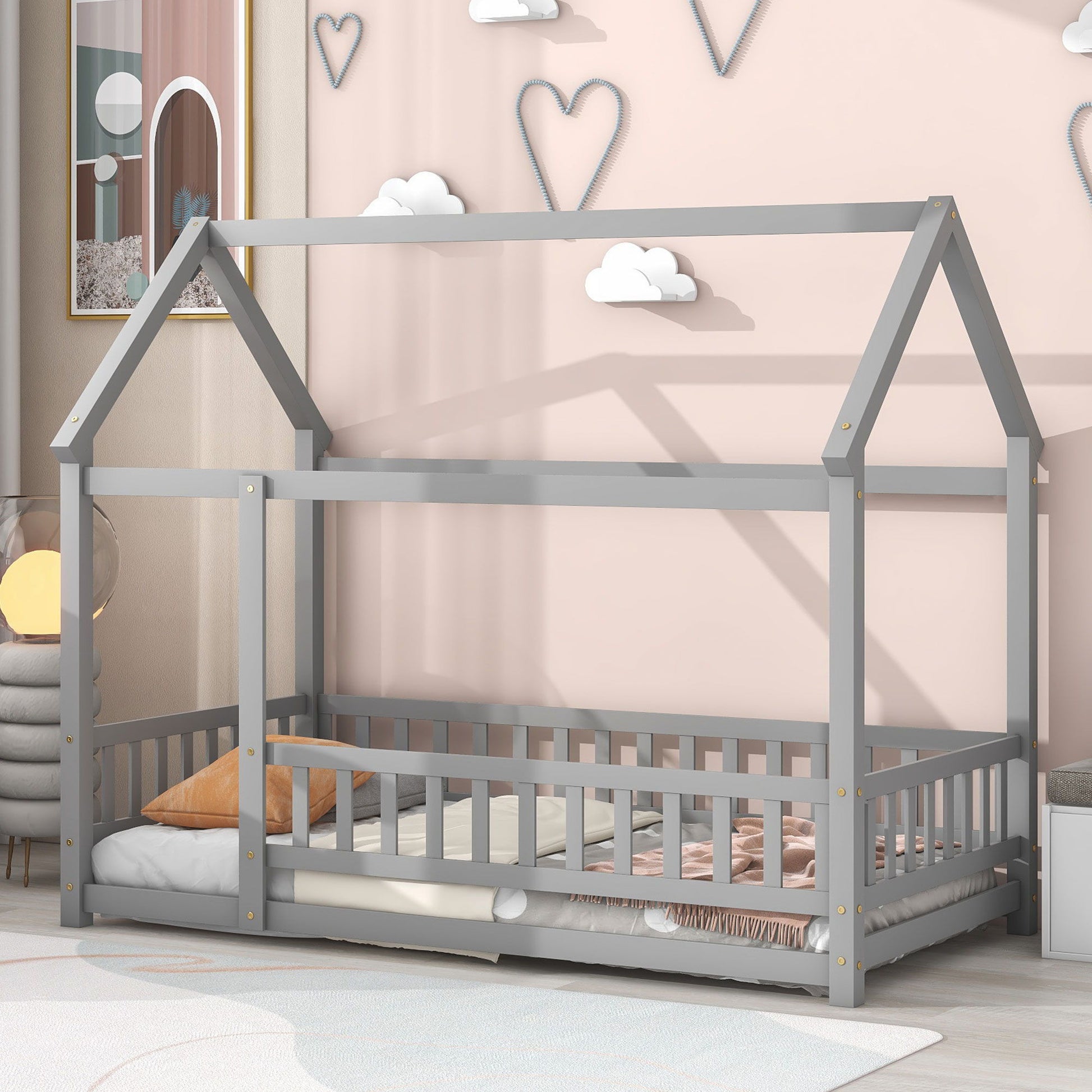 A Floor Wooden Bed With House Roof Frame and fence guardrails sits in a child’s room with pink walls decorated with hearts and clouds. The bed is styled with a white mattress, blush blanket, and orange pillow.
