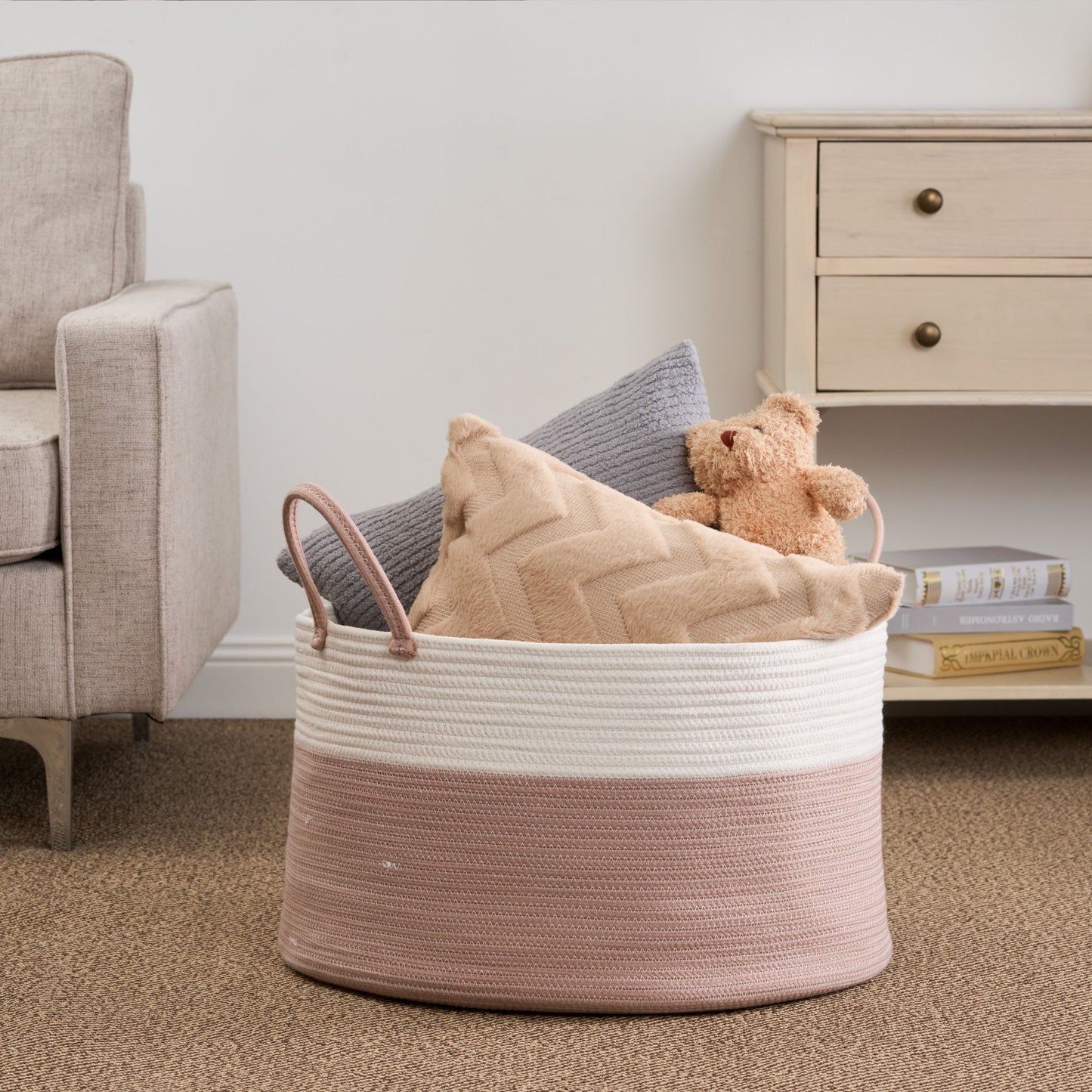 The Francis - Cotton Round Laundry And Storage Basket in pink and white sits on a carpeted floor, holding a gray pillow, a beige chevron pillow, and a tan teddy bear beside a beige armchair and light wooden dresser with books underneath.