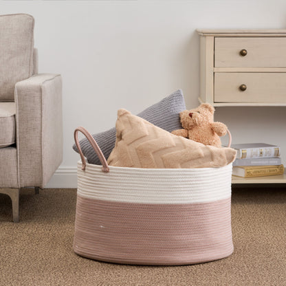 The Francis - Cotton Round Laundry And Storage Basket in pink and white sits on a carpeted floor, holding a gray pillow, a beige chevron pillow, and a tan teddy bear beside a beige armchair and light wooden dresser with books underneath.