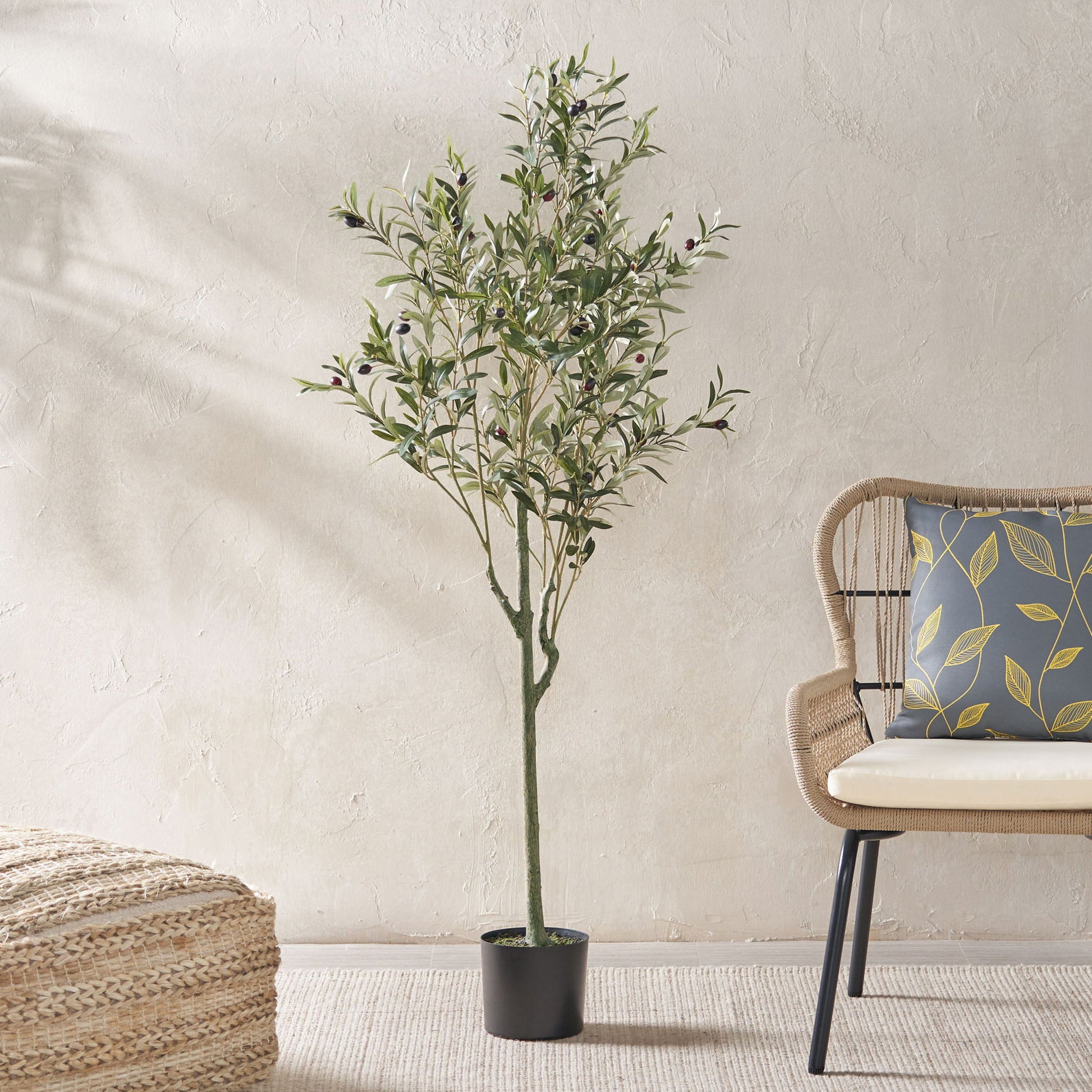 The Artificial Olive Tree with green leaves stands beside a wicker chair with a cushion and a woven ottoman in a light, minimalist room featuring a textured wall.