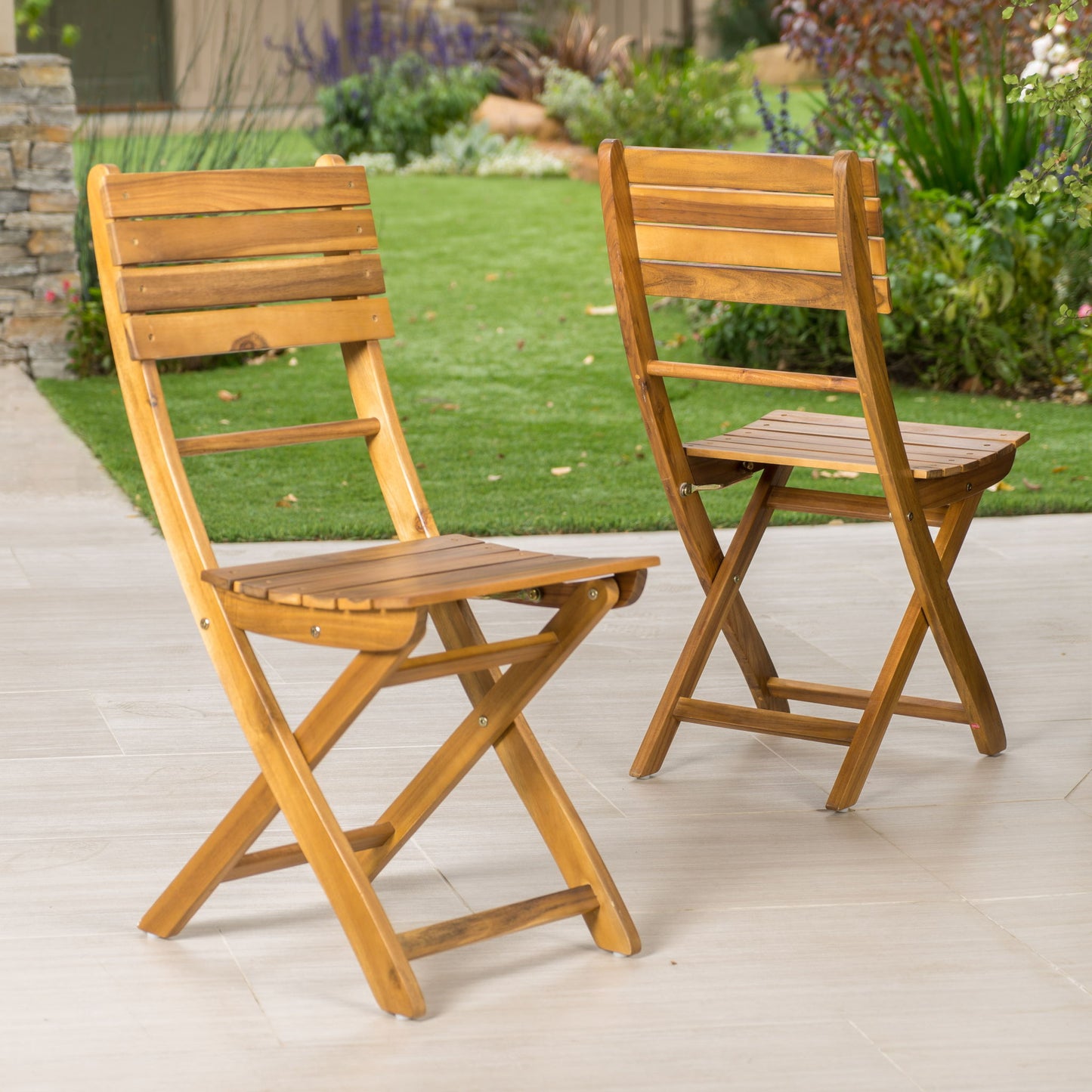 A set of two Outdoor Foldable Wood Dining Chairs with a slat design is arranged on a light patio, with a green lawn and garden plants in the background.