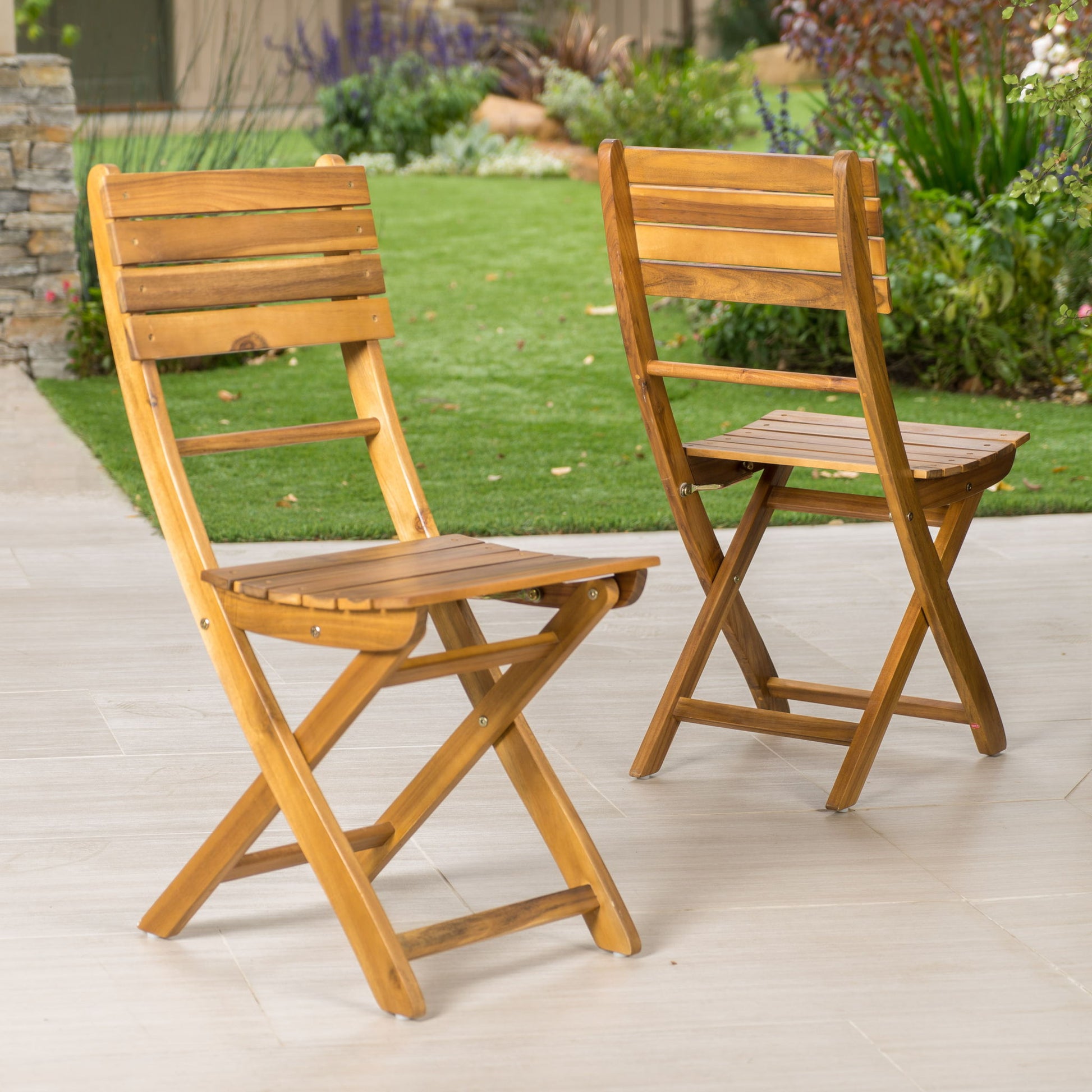 A set of two Outdoor Foldable Wood Dining Chairs with a slat design is arranged on a light patio, with a green lawn and garden plants in the background.