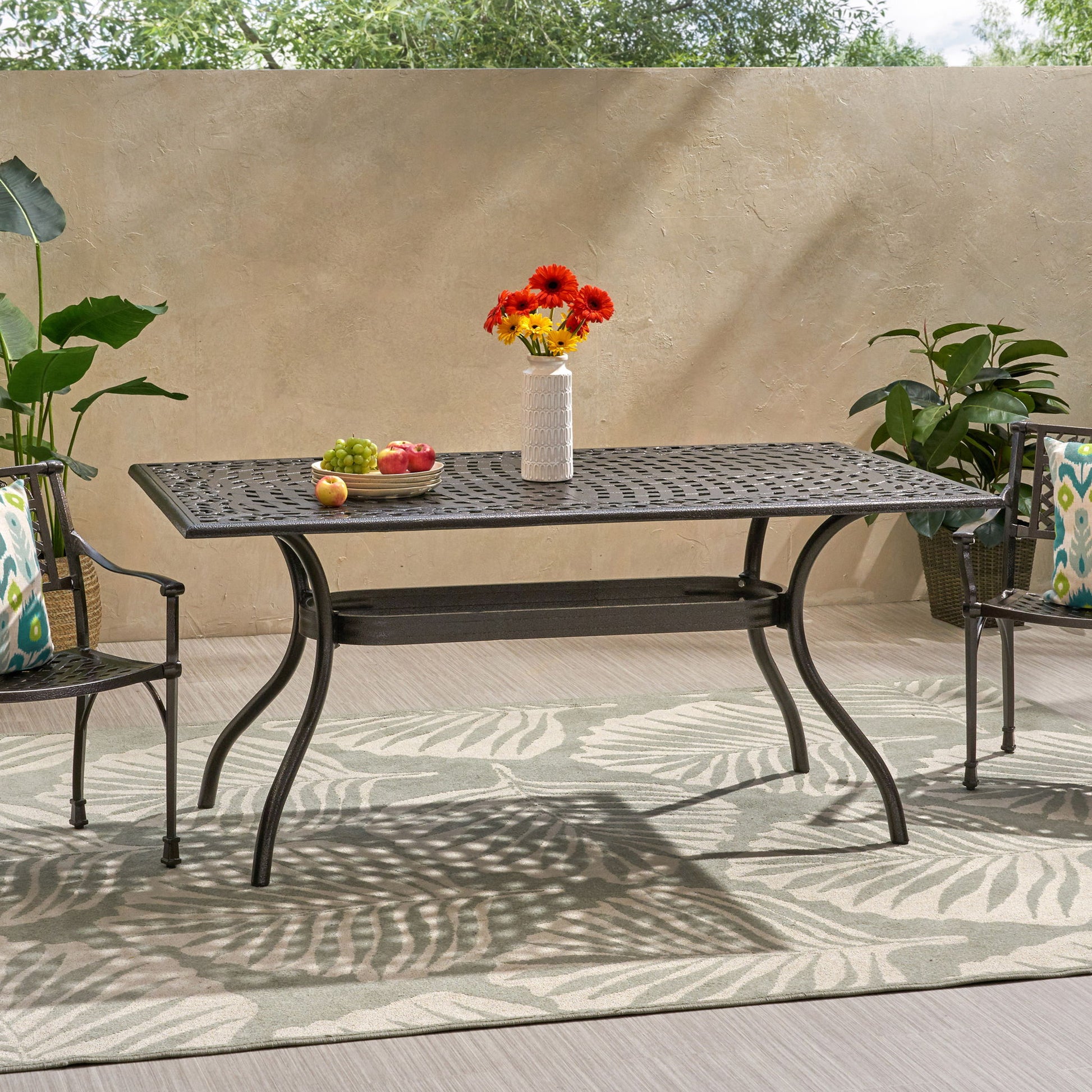 The Celestine Outdoor Cast Aluminum Dining Table with a floral lattice design and umbrella holder sits between two patterned chairs on a leafy rug, surrounded by potted plants against a beige wall.