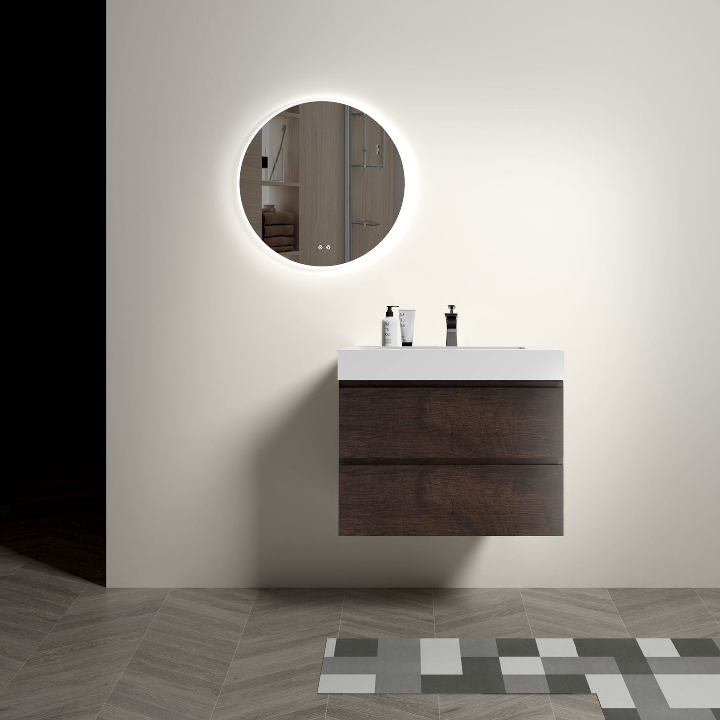 A minimalist bathroom features the Alice wall-mounted floating vanity with a one-piece sink basin, dark wood drawers, and a backlit round mirror. The white countertop holds two containers and geometric tiles cover the floor.