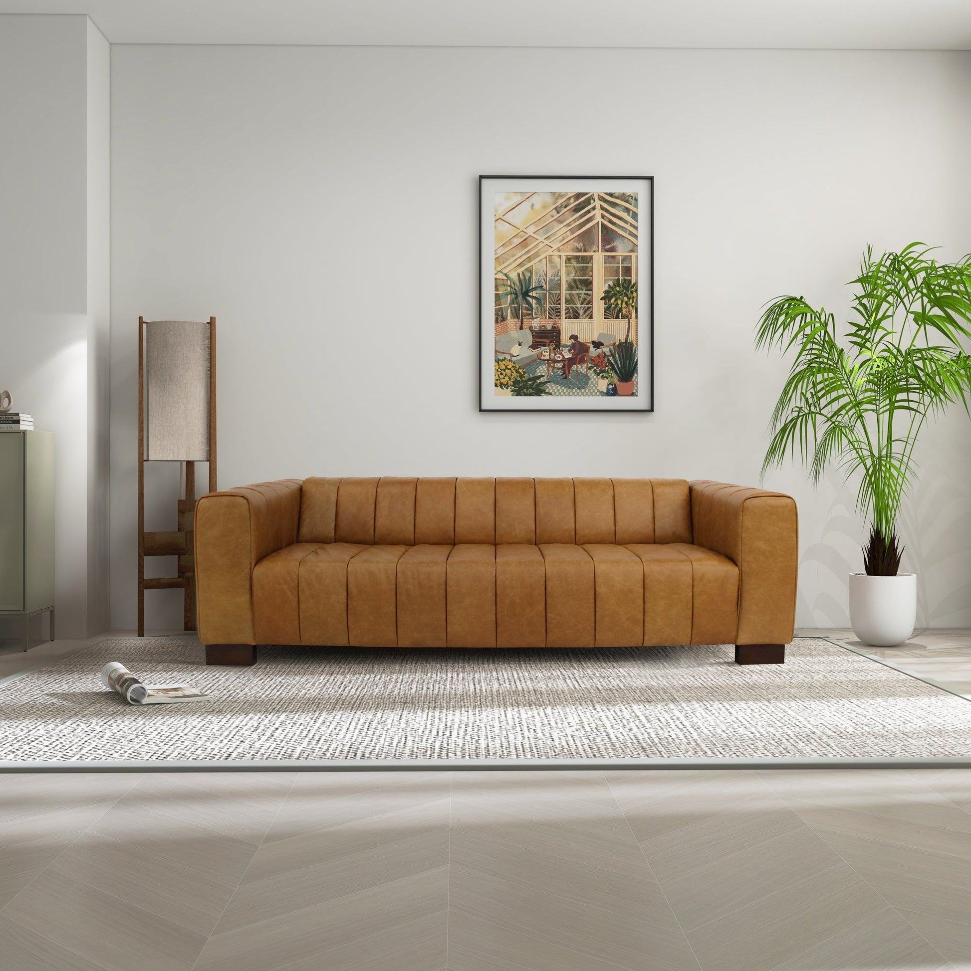A modern living room features the Dixon - Waxy Sofa - Tan on a light rug, paired with a potted palm, standing lamp, and framed greenhouse artwork on white walls. A rolled-up magazine rests on the rug.
