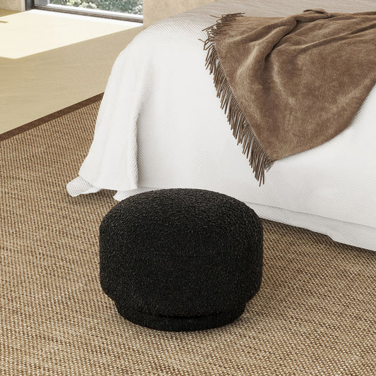 The Fuji - Mushroom Footstool Ottoman, round and black with a textured finish, sits on a woven rug beside a bed adorned with a white blanket and brown throw in a softly lit modern bedroom.