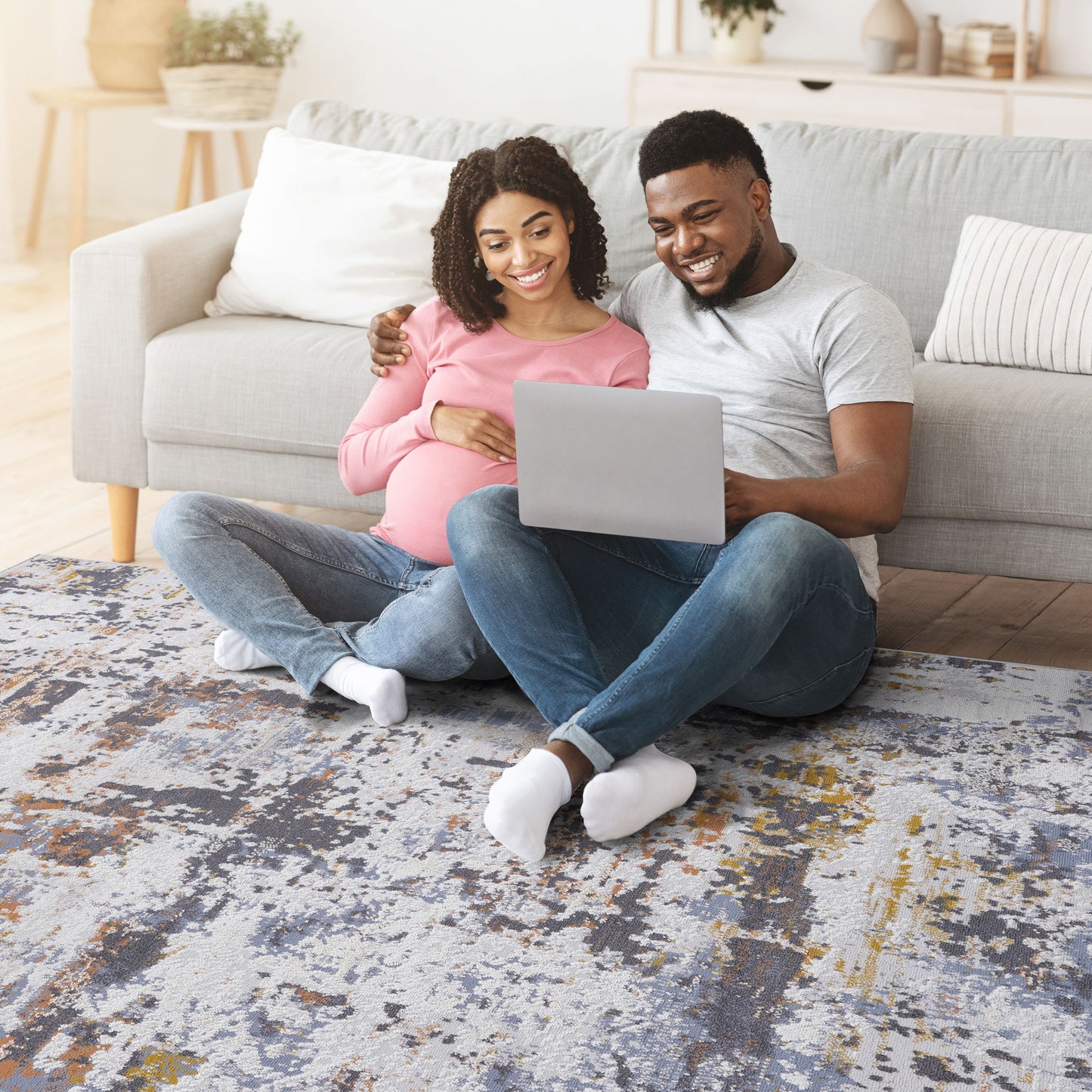 A happy pregnant couple sits on the floor by their sofa, smiling at a laptop together. The cozy scene is complete with the stylish Payas - Abstract Area Rug beneath them, creating a relaxed atmosphere at home.